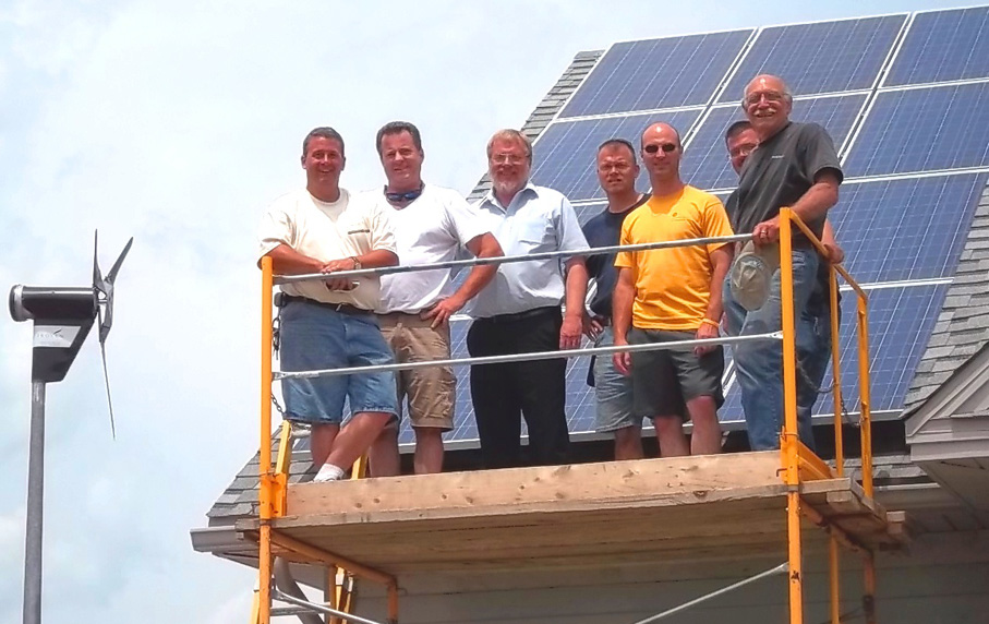 Workshop instructors and participants on scaffolding in front of solar panel with wind turbine to the left of them.